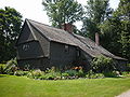 A large brown colonial-style house with multiple leanto additions. There is a flower garden in front of the house.