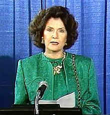 Woman in a green suit with a large gold necklace standing behind a podium