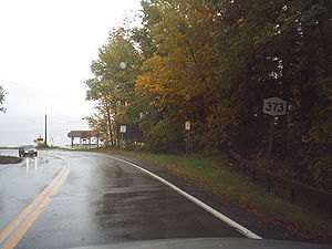 A two-lane highway turns to the left as it approaches a body of water. To the right of the highway is a NY 373 shield.