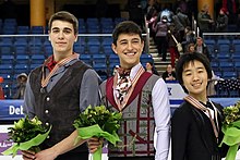 Die drei Gewinner stehen nebeneinander (Nadeau links) auf der Eisfläche. Alle drei halten Blumensträuße in der Hand und tragen ihre Medaillen an Bändern um den Hals. Im Hintergrund sieht man die sich leerende Zuschauertribüne.