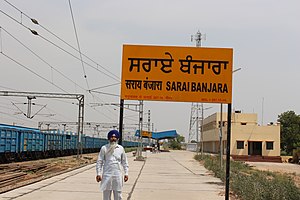 A man standing in front of an orange sign written in Hindi and English, with the text "Sarai Banjara". In the background a railway station.