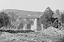 Foto hitam putih dari sebuah jembatan, dengan duduk sentry di kanan dan sebuah benteng di puncak ketinggian di kiri atas.