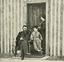 Photographie de l'entrée d'une maison en bois où un homme barbu en uniforme militaire est assis sur un fauteuil. Un garçon se tient à ses cotés et une femme en robe se trouve derrière eux.