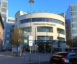 Three floor beige and circular building with big glass windows. Seen from the front, a Main Entrance metallic sign is above the door, and between the second and third floor, "University Hospital" is displayed with big letters.