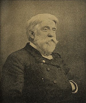 A white-haired man with a beard and mustache, facing left. He is wearing a white shirt, black vest and black jacket