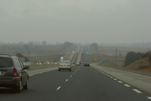 "A photograph taken driving down a highway on a foggy day. The highways passes over several humps in a roller-coaster like fashion before disappearing over one hump. It is straight throughout the photo. The setting is rural, with some trees interspersed among large fields, resembling a classic interpretation of the English countryside."