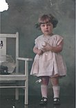 Photographie colorisée. Janet Rosenberg, enfant, posant en robe lors d’une séance de photographie. À gauche, un aquarium est posé sur une chaise.