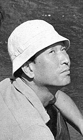 Homme asiatique avec un chapeau de type bob, photographié en plongée et de trois quarts, le regard tourné vers le haut.