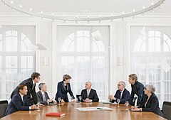 Huit personnes autour d'une table. Des papiers volent au dessus d'eux. Sur la table se trouvent la constitution de la Suisse et une carte de la Suisse avec une boussole.