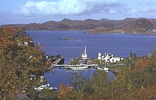 Un petit port entouré d'arbres abrite plusieurs yachts, bateaux de pêche et une vedette à moteur.  Le port est relié à la mer au-delà par une porte d'écluse.  Un petit rocher se trouve dans l'eau et des collines rocheuses brunes dominent au loin.