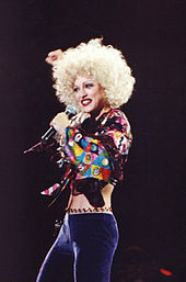 Image of a blond female facing her left. She has short blonde hair and is wearing a green bra and purple pants with beads on the waist. She is singing to a microphone, which she holds to her mouth with her left arm.