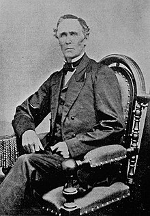 An old, frowning man wearing a suit sitting in a chair with his right arm (left from the perspective) leaning on the arm of the chair