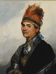 A head and shoulders oil portrait of Joseph Brant. He wears Indian garb, including a wide headband decorated with feathers and beads, a metal gorget, and a dark cape with a silver fringe.