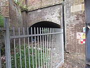 A tall brown brick wall has a tunnel entering it. The tunnel is approximately 20 metres long & 3 metres wide, going to some green foliage. In front of the tunnel is a metal fence. To the right of the image are some danger signs.