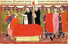 Um homem com coroa deitado na cama recebe a Eucaristia de dois sacerdotes