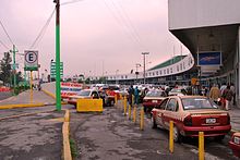 Picture of the main entrance to the Northern Bus Terminal. Multiple cars are parked and many pedestrians walk on the sidewalk.