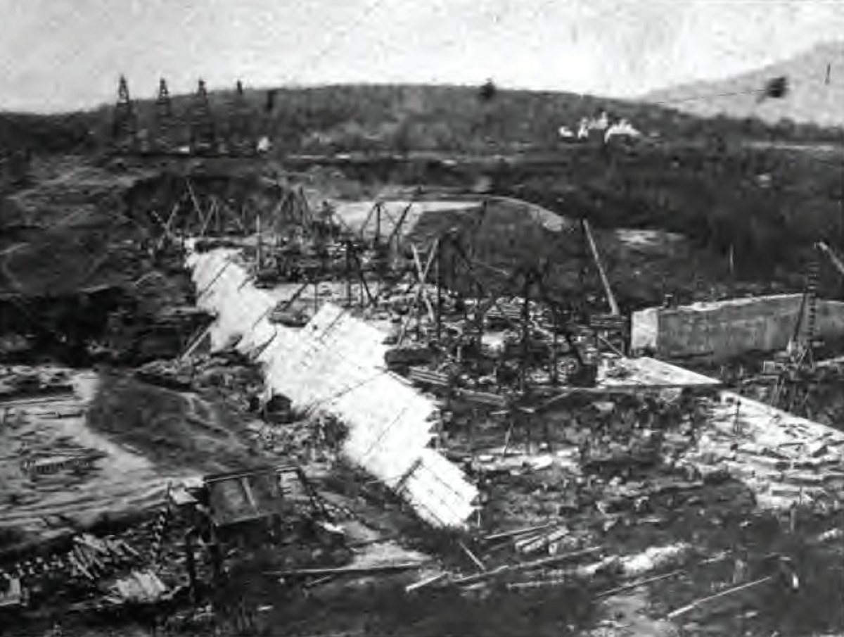 A black and white photograph of the lower sections of a sloped dam， with cranes and other construction equipment on top