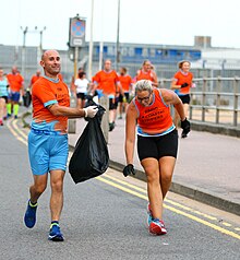 A woman bending to pick up litter while a man jogs alongside, with a garbage bag, at a plogging event in Kent, England Plogging with Coastal Striders - FUNK9730 (42405061590).jpg