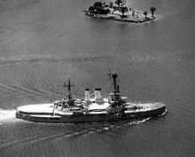 A large warship, its decks covered in canvas shades, creates large waves as it sails through shallow water. On its left side, it passes a small island with several trees and a small building.