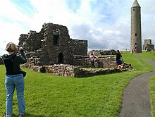 Fothrach agus daoine á thaiscéal. Tá bean chun tosaigh sa phictiúr ag glacadh pictiúr den fhothrach. Tá cloigtheach chun cúl.