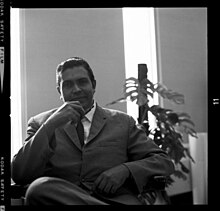 Rienzi Crusz wearing a suit and seated in an armchair looking toward the camera