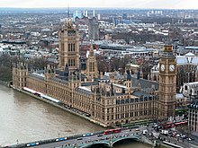 Large sand-coloured building of Gothic design beside brown river and road bridge. The building has several large towers, including large clock-tower.