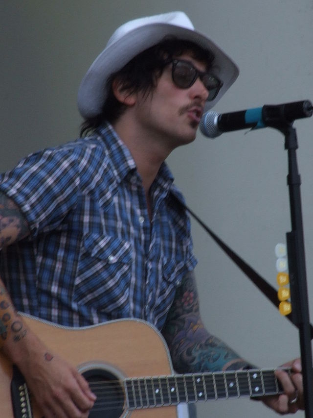 A man wearing a cowboy hat and sunglasses playing a guitar in front of a microphone stand.