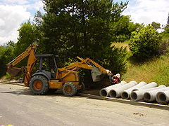 A worker attaches a lifting cable to a concrete sewer pipe section. Note the retracted stabilizers on this Case backhoe.