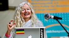 Jutta Hartmann ist mit vor einem unscharfen Hintergrund zu sehen, während sie vor einem Laptop sitzt und den rechten Arm halb erhoben zu einer Geste positioniert hat. Sie ist während des Sprechens zu sehen und hat lange helle gelockte Haare.