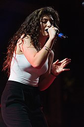 Lorde wearing a white crop top and black trousers singing onstage while closing her eyes