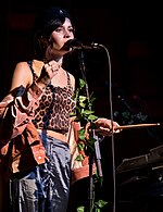 A woman with dark hair, a crop top and satin pants stands in front of a microphone stand