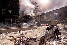 New York fire department personnel examining a smashed New York City police car, during 9/11 Rescue worker reaching into a New York Police car covered with debris (28802606564).jpg