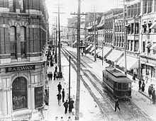 Black and white image of a downtown Ottawa street scene circa 1909, including an electric tram travelling down tracks on the street, rows of buildings on both sides of the street, the railway office building on a street corner, and numerous pedestrians on both sidewalks