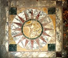 The floor mosaic made of heavily marbled stone slabs shows a symbol of the sun: In the middle an ocher-colored sun disk with a thin black border, starting with 16 rust-red rays (pointed triangles), embedded in an outer white disk.  This sun disk is in turn surrounded by an ocher-colored square with four smaller black squares in the four corners.