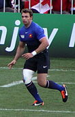 De trois-quarts, un homme au maillot bleu, le genou droit bandé, court sur un terrain de rugby.