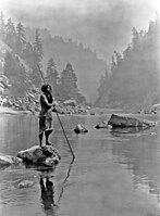 A smoky day at the Sugar Bowl—Hupa, c. 1923. Hupa man with spear, standing on rock midstream, in background, fog partially obscures trees on mountainsides.
