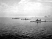 Sebuah foto hitam dan putih dari empat kapal-kapal besar berlayar di laut tenang dari kanan ke kiri.