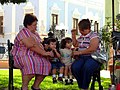 Two young children sit between two women on a park bench in Campeche City in 2006. The children are drinking beverages through straws that they hold. The children are looking away from the beverage containers, which the women are holding for them.