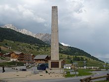 Farbfoto eines großen Obelisken am Ende eines Dorfes vor den Bergen