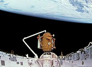 A space shuttle payload bay, covered in white insulation, with a small, cylindrical orange module at one end, supported by the shuttle's robotic arm. The blackness of space and the Earth serve as the backdrop.