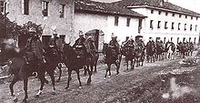 A line of horses ridden by men in military uniforms. The horses are moving along a dirt road that runs alongside several multi-story buildings.