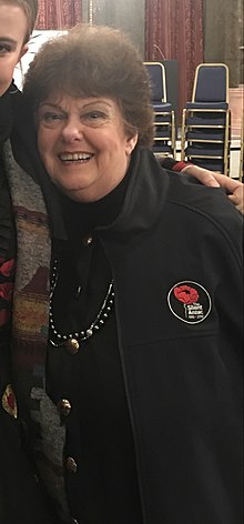 Margaret Haggart standing looking at camera smiling, she is wearing a jacket with the label "The Silent Anzac" behind her a stacked chairs and marble columns.