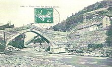 Photographie en noir et blanc d'un pont sur l'Arc en 1930 à Orelle, sur une carte postale au musée municipal, enjambant la rivière de l'Arc et présent à côté de maisons d'époque.