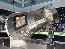 A conical, metallic-grey spacecraft, with a hole cut in one side to allow access, on a display stand inside a museum. It is covered in a close-fitting transparent plastic sheath.