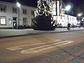 Station forecourt in Marburg with a white elongated 20 painted on the asphalt road surface.  Main entrance in the evening at Christmas time with illuminated Christmas tree (December 27, 2016).  The area marked off by a curb directly in front of the stairs to the entrance to Marburg Central Station (left) is reserved for pedestrians and pushing cyclists, a zero lowering in the curb (curb lowering) also makes it easier for wheelchair users to access, with a bright, tactile attention field at the roadside “Also blind the beginning of the road;  In addition to city buses, the area at the bus station can also be used by bicycles, and the Neue Kasseler Strasse, which continues to the left, is an important bicycle route.  Some bicycles are parked on the house wall behind the Christmas tree near the brightly lit entrance, but not directly in front of the stairs, a little further to the left outside of the picture there is a covered storage area for bicycles.