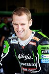 A man in his late twenties is smiling and wearing green and black motorcycling overalls