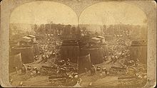 Dos fotos idénticas de una calle, atestada de varios carros tirados por caballos, con grandes tanques de almacenamiento a cada lado y torres de perforación de petróleo visibles en la distancia.  Las imágenes están montadas una al lado de la otra en una tarjeta.