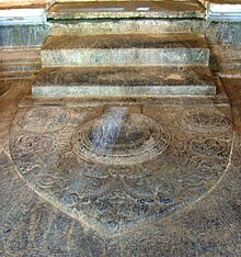 Spitzflachstein;  in der Mitte ein runder Lotus im Relief und zwei kleine an den Seiten, das Ganze auf dem Laubboden.