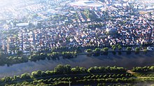 Luftbild des Flörsheimer Stadtzentrums. Im Vordergrund der Main mit der katholischen Kirche St. Gallus (mit Reflexion) dahinter. Im Hintergrund das Einkaufszentrum und die weiterführende Schule, 2015