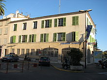 Un long bâtiment de style provençal avec des barreaux aux ouvertures du rez-de-chaussée et des volets verts aux deux étages.